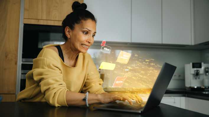 Eine Frau schaut besorgt auf einen Laptop, während ihr Hassbotschaften entgegenfliegen (Bild: picture alliance / Westend61 | Infinite Lux)