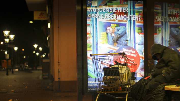 Ein Mann übernachtet in einer Winternacht bei Minusgraden an einer Bushaltestelle in Berlin-Schöneberg, dahinter eine Werbetafel der Berliner Stadtmission mit einem Spendenaufruf für ihr Kältehilfe-Projekt für Wohnungslose