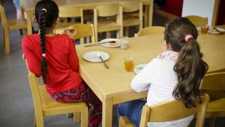 Zwei Kinder sitzen am Tisch und bekommen Essen von einer Schultafel