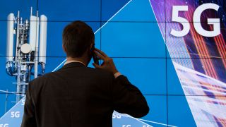 Ein Mann steht im Gebäude der Bundesnetzagentur mit Handy am Ohr vor einer Leinwand mit der Aufschrift "5G". (Bild: dpa)