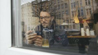 Ein Mann sitzt mit seinem Handy hinter der Fensterscheibe eines Berliner Cafés (Bild: imago/Westend61)