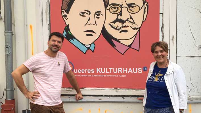 Kriss Rudolph und Christiane Härdel stehen vor dem Logo des Queeren Kulturhauses, bestehend aus der feministischen Schriftstellerin Johanna Elberskirchen und dem Sexualwissenschaftler Magnus Hirschfeld.