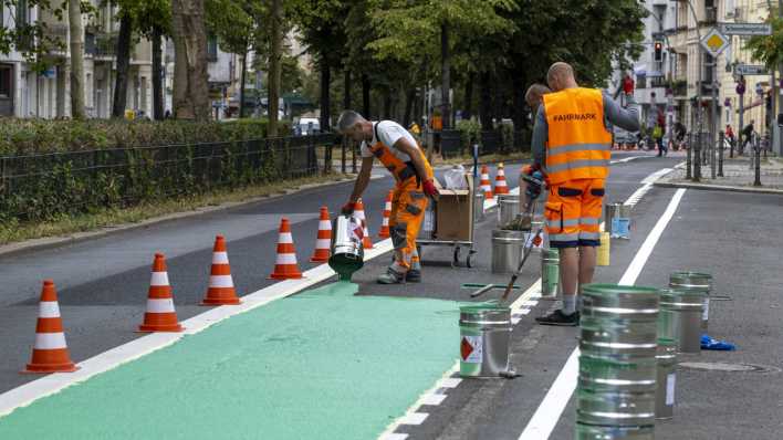 Bauarbeiter gießen mit grüner Farbe eine neue Fahrbahnmarkierung für einen Radweg (Bild: imago images / snapshot)