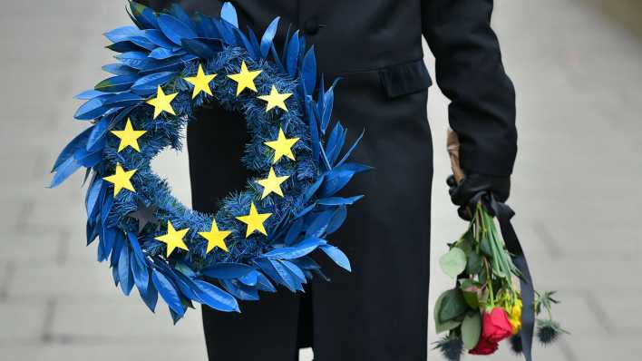 Ein Mann verkleidet als Bestatter hält einen Kranz in der Farben und Symbolen der EU
