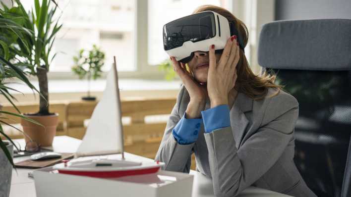 Symbolbild virtuelles Reisen: Frau mit VR Brille und Segelboot-Modell auf dem Schreibtisch (Bild: imago images / Westend61)