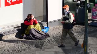 Ein Obdachloser sitzt an einer Einkaufsstrasse in Steglitz vor einem Laden und trägt einen Mundschutz. Ein älterer Mann mit Mundschutz geht an ihm vorbei (Bild: dpa)