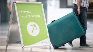 Ein Urlauber läuft mit seinem Koffer an einem Schild mit der Aufschrift "Corona-Testzentrum" vorbei. (Bild: dpa)