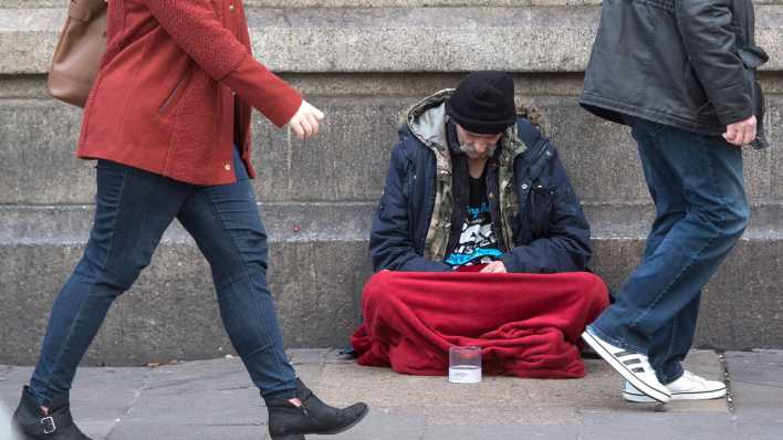 Ein obdachloser Mann sitzt mit gesenktem Kopf auf der Straße - Passanten laufen an ihm vorbei (Bild: dpa)