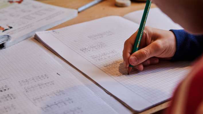 Ein Grundschüler sitzt zuhause in der Küche und rechnet in seinem Schulheft
