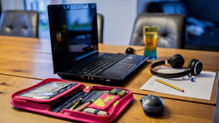 Auf einem Tisch in einem Wohnzimmer steht ein aufgeklappter Laptop, daneben liegt ein Federmäppchen, davor liegen Kopfhörer, ein Bleistift und ein Zettel (Bild: IMAGO / Fotostand)