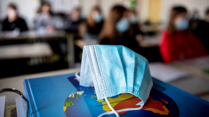 Eine medizinische Maske liegt auf einem Schulbuch. Im Hintergrund ist unscharf eine Schulklasse zu sehen, die ebenfalls Masken tragen (Bild: dpa / Matthias Balk)