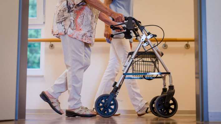 Eine Pflegekraft geht in einem Pflegeheim mit einer älteren Frau über einen Korridor. (Bild: dpa)