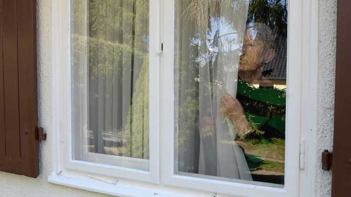 Eine ältere Frau steht hinter der Gardine am Fenster.