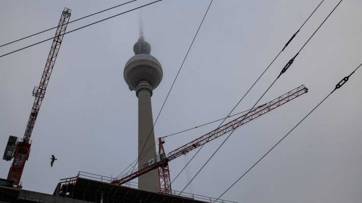 Die Spitze des Berliner Fernsehturms am Alexanderplatz im Bezirk Mitte verschwindet im Nebel waehrend im Vordergrund eine Baustelle und Baukraene zu sehen sind.