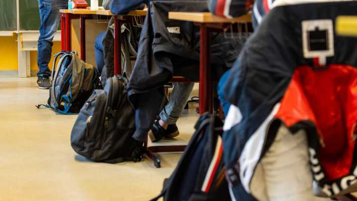 Rucksäcke stehen neben den Tischen, an denen die Schülerinnen und Schüler beim Unterricht in ihrer Klasse sitzen, während ein Lehrer vor der Tafel steht.