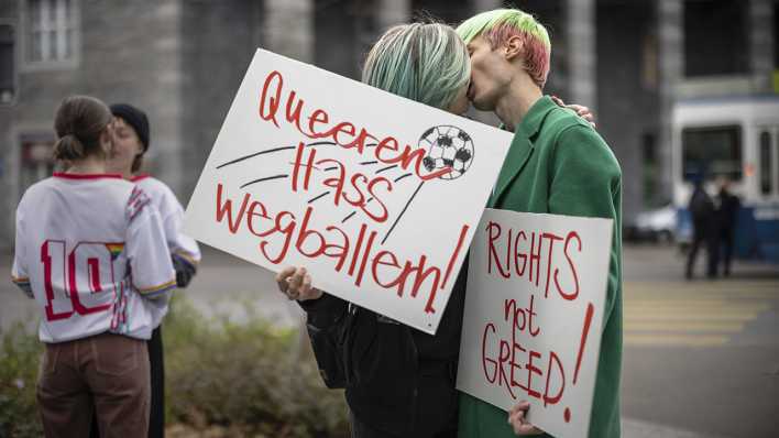 Demonstranten küssen sich aus Protest gegen die Fußball-WM in Katar. Sie halten ein Schild mit der Aufschrift "Queeren Hass wegballern!" in der Hand.