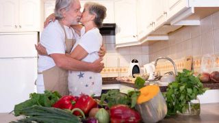 Gesunde Ernährung im Alter: Paar küsst sich in Küche, im Vordergrund Gemüse (Bild: imago images/Westend61)
