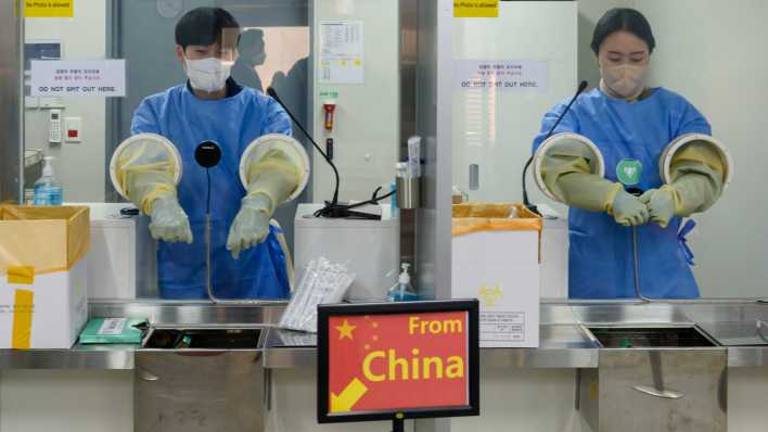 Zwei Mitarbeiter machen sich an einem Flughafen bereit für einen Corona-Test, davor ein Schild mit der Aufschrift "From China".