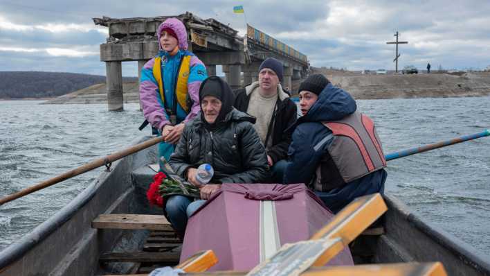 Vier Menschen überqueren auf einem Boot einen Fluss, vor ihnen liegt ein Sarg, im Hintergrund ist die ukranische Flagge zu sehen.