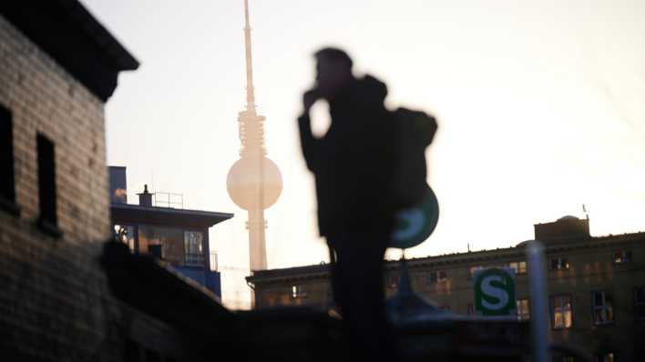 Die Sonne geht hier dem Fernsehturm auf, während ein Mann mit Rucksack am Nordbahnhof im Gegenlicht steht und telefoniert.