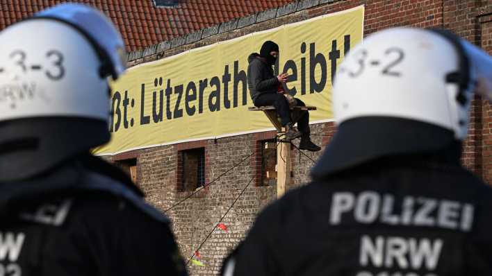 Ein Demonstrant sitzt bei der Räumung des Dorfes Lützerath auf einer Holzkonstruktion vor einem Plakat mit dem Slogan "Lützerath bleibt!" Im Vordergrund sind polizisten zu sehen. (Quelle: dpa)