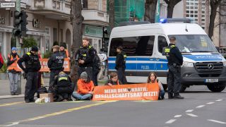 Aktivisten der Gruppe "Letzte Generation" haben sich auf der Neuen Kantstraße auf einer Kreuzung festgeklebt. Die Polizei sichert den Bereich.