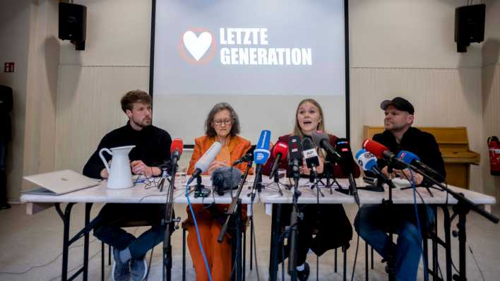 Aktivisten der "Letzten Generation" sitzen bei einer Pressekonferenz nach den Razzien gegen ihre Gruppe an einem Tisch voller Mikrofone (Bild: dpa)