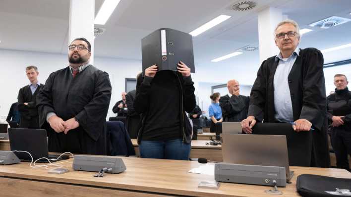 Die Angeklagte Lina E. (M) steht bei der Fortsetzung des Prozesses im Oberlandesgericht (OLG) Dresden im Verhandlungssaal und hält einen Aktenordner vor ihr Gesicht. (Bild: dpa)