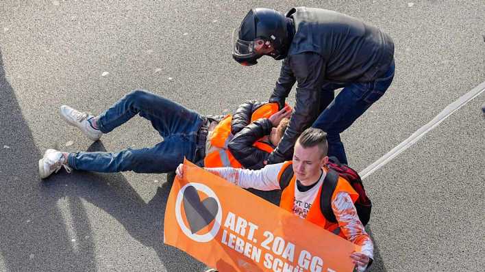 Die Klimaaktivisten blockieren mit Bannern die Fahrbahn, einige haben sich festgeklebt. Aufgebrachte Verkehrsteilnehmer, die durch die Aktivisten aufgehalten werden greifen ein und versuchen die Protestler von der Straße zu holen.