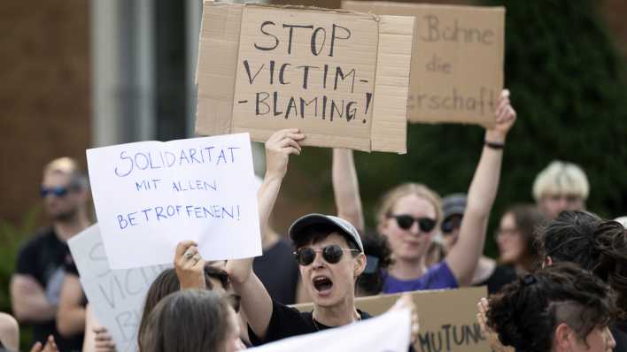 Menschen demonstrieren gegen einen Auftritt von Rammstein und halten Plakate mit der Aufschrift "Stop victim blaming" und "Solidarität mit allen Betroffenen" hoch.