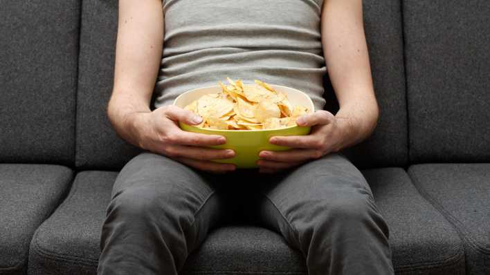 Ein Mann sitzt auf dem Sofa und hält eine Schüssel mit Chips auf dem Schoß.