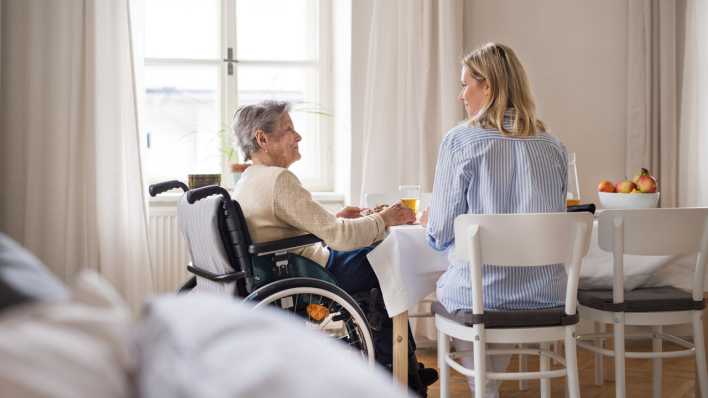 Eine jüngere Frau und eine ältere Frau im Rollstuhl sitzen gemeinsam an einem Esstisch.