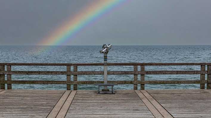 Symbolfoto Zukunftsangst: Ein Fernglas vor einem Meer mit dunklen Wolken und einem Regenbogen.