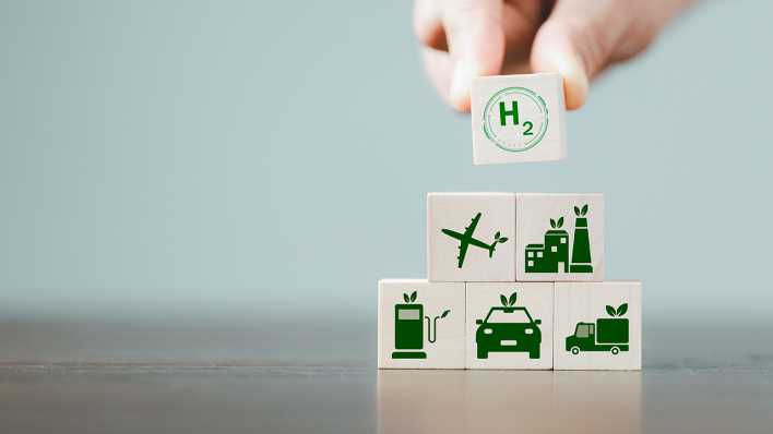 Jemand stapelt Würfel übereinander: darauf sind ein Flugzeug, eine Fabrik, Autos und H2, das Zeichen für Wasserstoff, zu sehen.