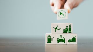 Jemand stapelt Würfel übereinander: darauf sind ein Flugzeug, eine Fabrik, Autos und H2, das Zeichen für Wasserstoff, zu sehen.
