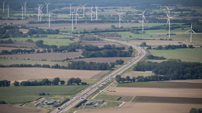 Autos und Lastwagen fahren auf der A24. Zu sehen ist auch der Rastplatz Ruppiner See sowie Windräder am Horizont.