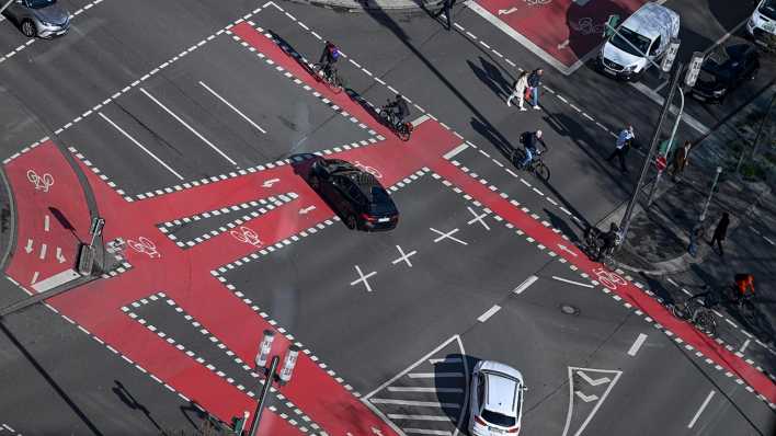 Radfahrer, Fußgänger und Autos sind an einer Kreuzung, die mit roten Fahrradspuren markiert ist, unterwegs.