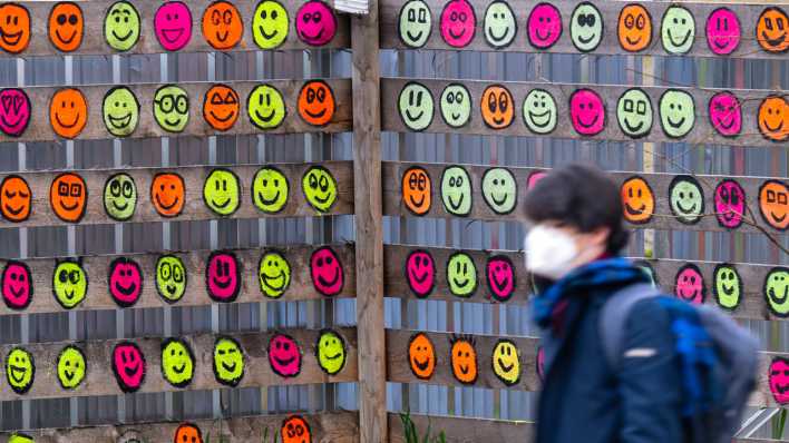 Eine Passantin mit Mund-Nasen-Bedeckung geht an einem hölzernen Bauzaun vorbei, der mit bunten Smileys bemalt wurde (Bild: dpa / Peter Kneffel)