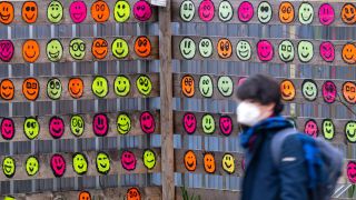Eine Passantin mit Mund-Nasen-Bedeckung geht an einem hölzernen Bauzaun vorbei, der mit bunten Smileys bemalt wurde (Bild: dpa / Peter Kneffel)