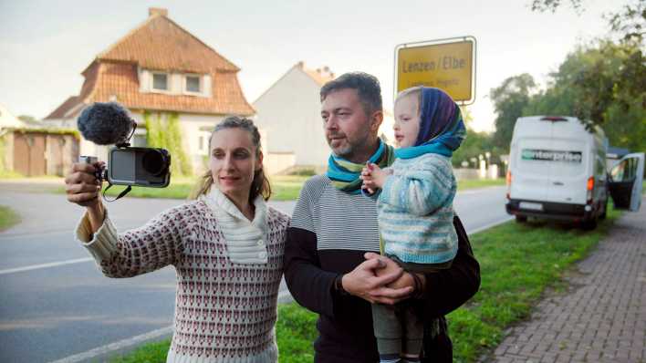Anne Münch mit Mann und Kind vor dem Ortseingangsschild von Lenzen in der rbb-Doku.
