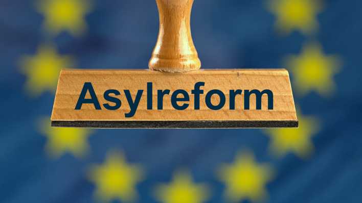 Ein symbolischer Holzstempel mit der Aufschrift "Asylreform", gehalten von einer Hand im Anschnitt vor einer unscharfen EU-Flagge im Hintergrund. (Bild: picture alliance / SULUPRESS.DE | Torsten Sukrow)