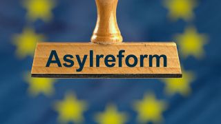 Ein symbolischer Holzstempel mit der Aufschrift "Asylreform", gehalten von einer Hand im Anschnitt vor einer unscharfen EU-Flagge im Hintergrund. (Bild: picture alliance / SULUPRESS.DE | Torsten Sukrow)