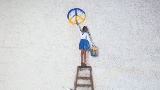Streetart auf einem Gebäude in Bucha in der Ukraine, das ein Mädchen zeigt, das ein Peace-Zeichen in ukrainischen Farben an eine Wand malt (Bild: picture alliance / AA | Oleksii Chumachenko)