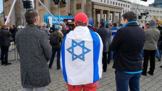 Auf der Kundgebung "Gegen Terror und Antisemitismus" in Berlin stehen drei Männer nebeneinander. Der Mann in der Mitte trägt eine Israel-Flagge um die Schultern (Bild: picture alliance / Marc Vorwerk)