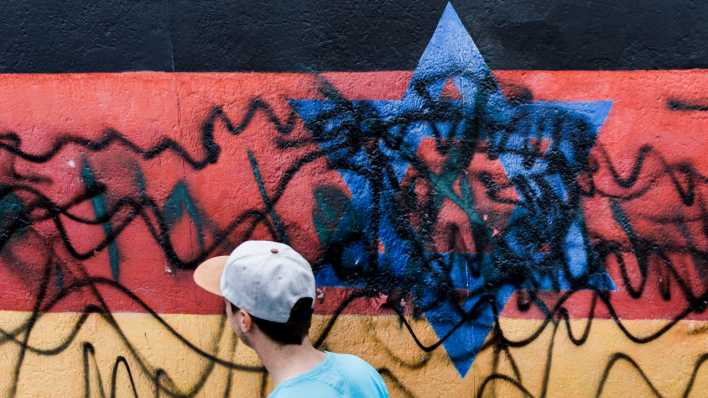 Ein junger Mann geht an dem zerstörten Wandbild "Vaterland" des Künstlers Günther Schäfer an der East Side Gallery vorbei. Das Wandbild, das die schwarz-rot-goldene Deutschlandfahne mit einem blauem Davidstern zeigt, wurde 2020 von Unbekannten mit schwarzen Graffitis beschmiert (Bild: dpa / Carsten Koall)