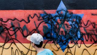 Ein junger Mann geht an dem zerstörten Wandbild "Vaterland" des Künstlers Günther Schäfer an der East Side Gallery vorbei. Das Wandbild, das die schwarz-rot-goldene Deutschlandfahne mit einem blauem Davidstern zeigt, wurde 2020 von Unbekannten mit schwarzen Graffitis beschmiert (Bild: dpa / Carsten Koall)