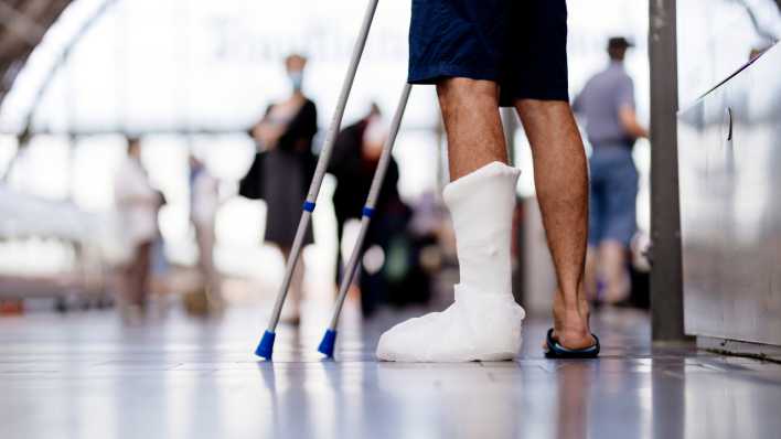 Symbolfoto Krankenversicherung: Ein Reisender mit Gipsbein steht am Bahnghof.