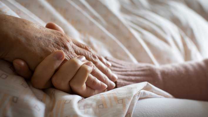 Symbolfoto: Eine Person hält am Bettrand die Hand einer älteren Person