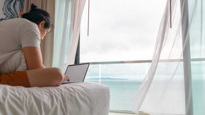 Ein Mann sitzt auf einem Bett und arbeitet an einem Laptop, im Hintergrund sieht man aus dem Fenster das Meer.
