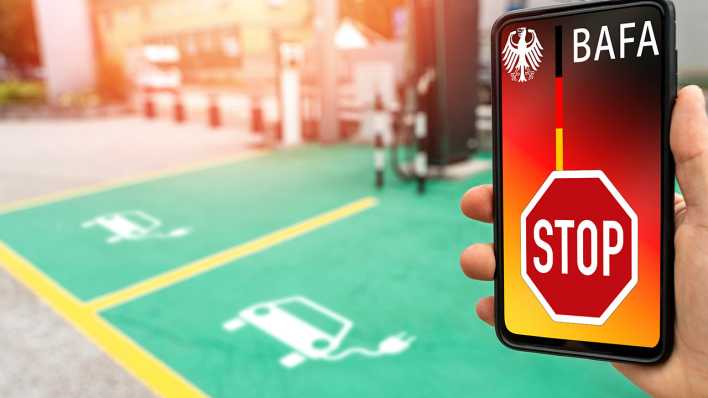 Ein Smartphone zeigt das Wort "BAFA", darunter ein Stopschild. Im Hintergrund ist ein Parkplatz für Elektro-Autos zu sehen, auf dem man das Auto abstellt, um dann die Batterie des Autos laden zu können.